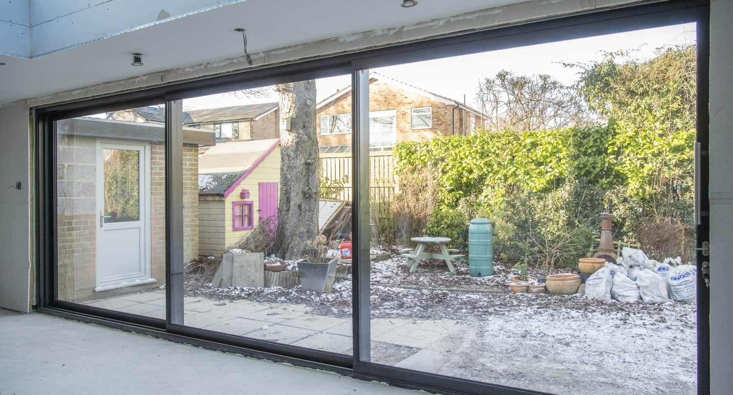 View through large bespoke glass sliding doors reveals a backyard with a tree, patio, shed, picnic bench, green water barrel, bags of material, and a thin layer of frost covering the ground and surfaces.