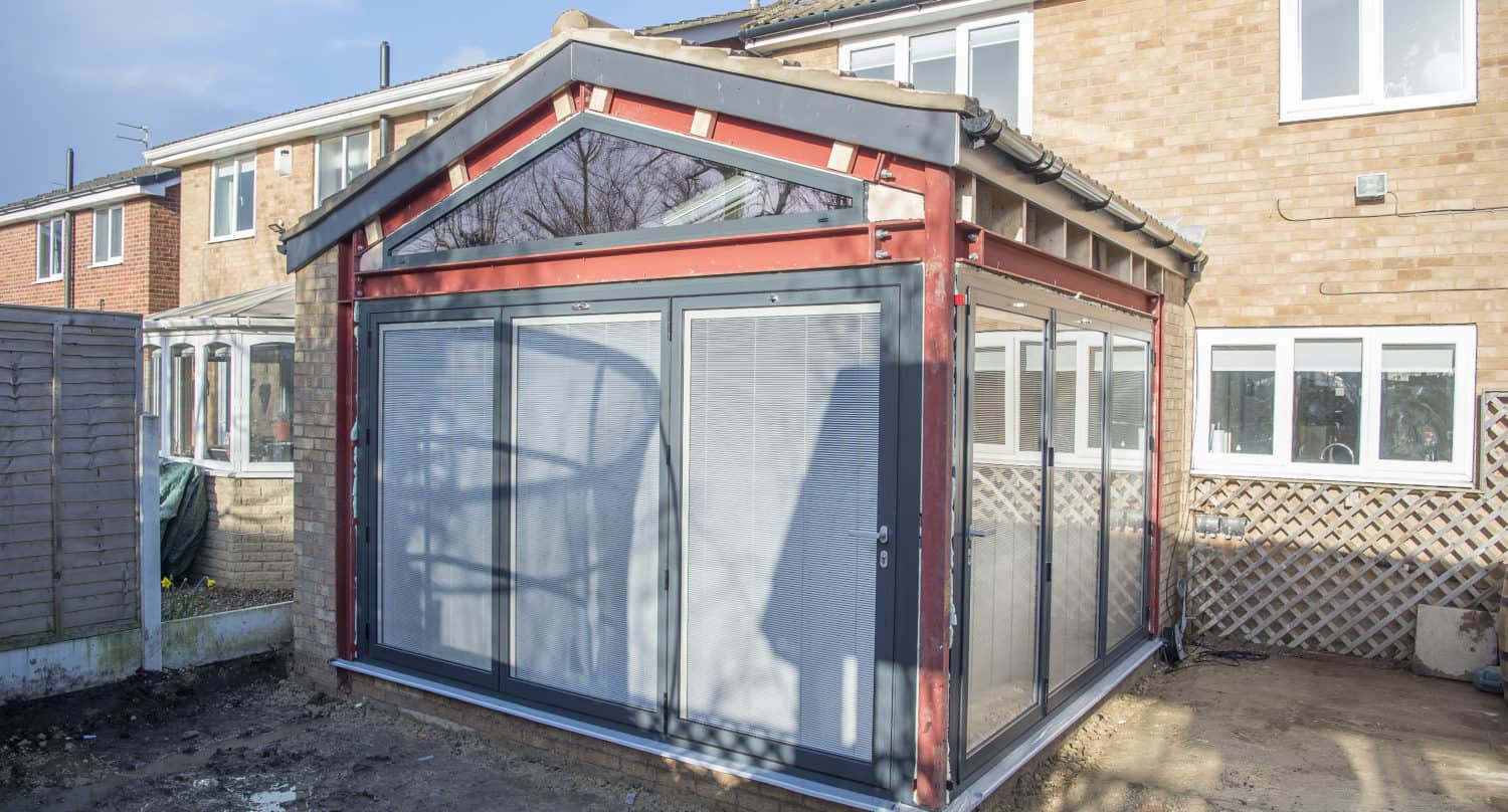 A modern house extension with a steel frame, large glass windows, bespoke glass solutions, and sliding doors is attached to the back of a brick house; the ground around is bare and unfinished.