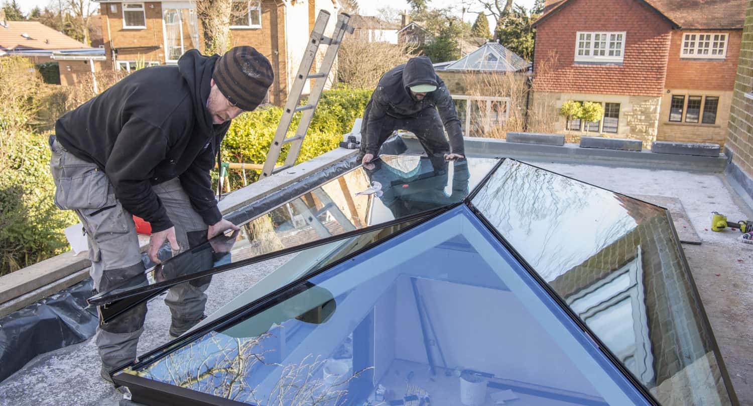 Two workers install glass panels on a modern rooftop skylight with glass roofing, using gloves and tools, while houses and trees are visible in the background on a clear day.