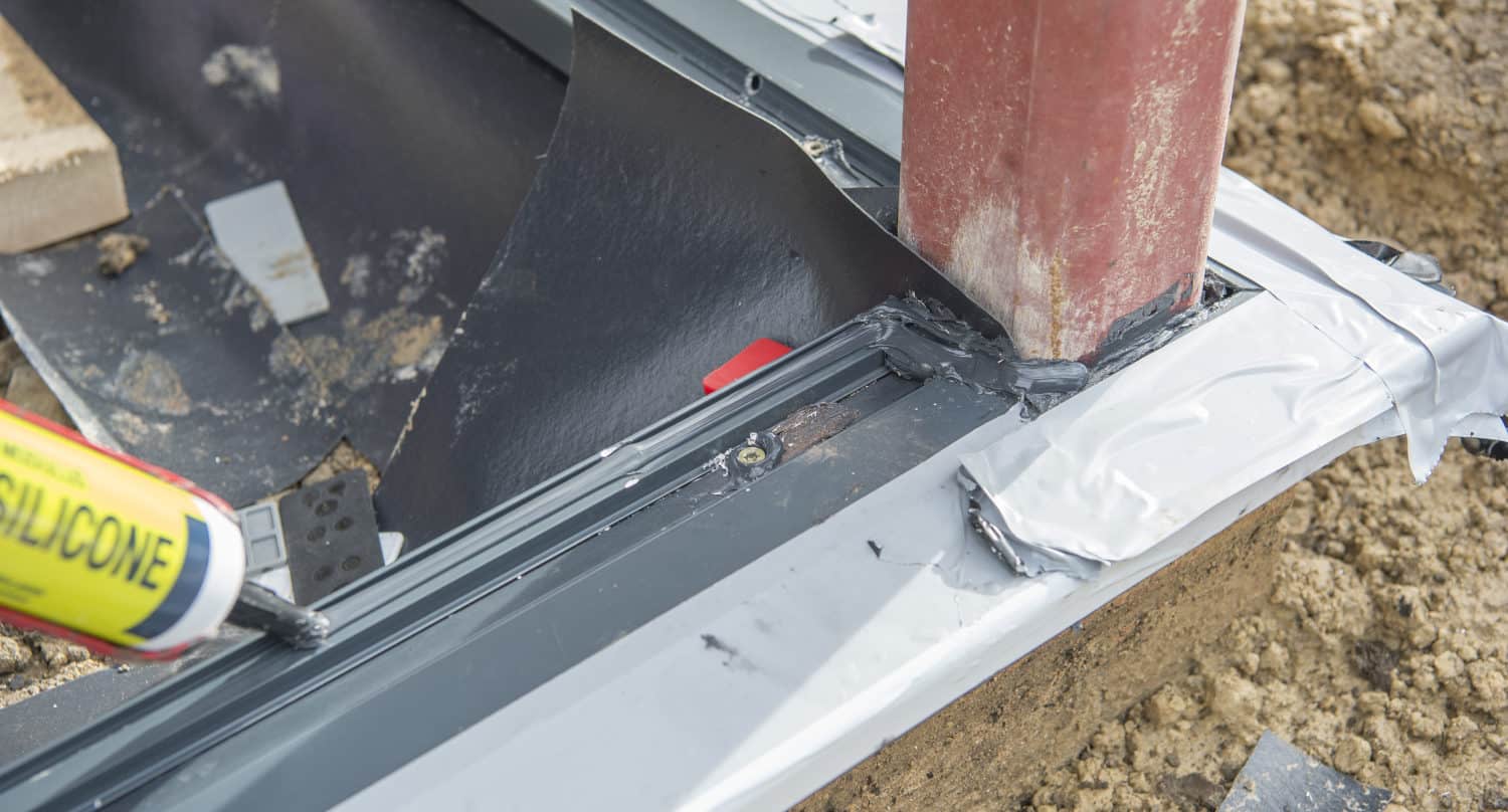 A close-up of a construction site shows a person applying sealant around the base of a metal post near glass roofing, with metal flashing and tape in place and soil visible in the background.