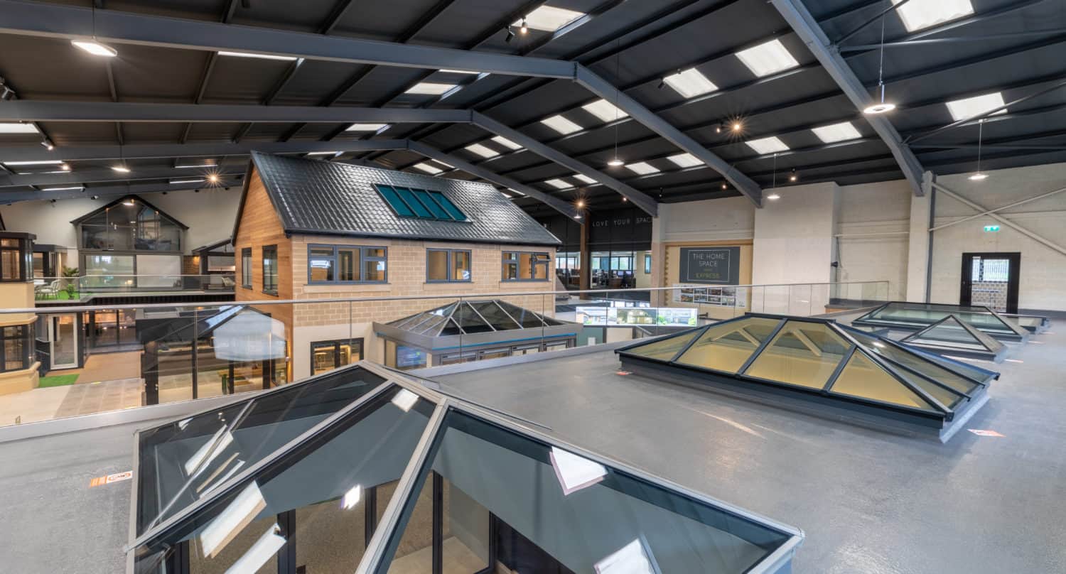 A spacious indoor showroom displays various house models, front doors, skylights, and windows under a high, industrial-style ceiling with bright lighting.