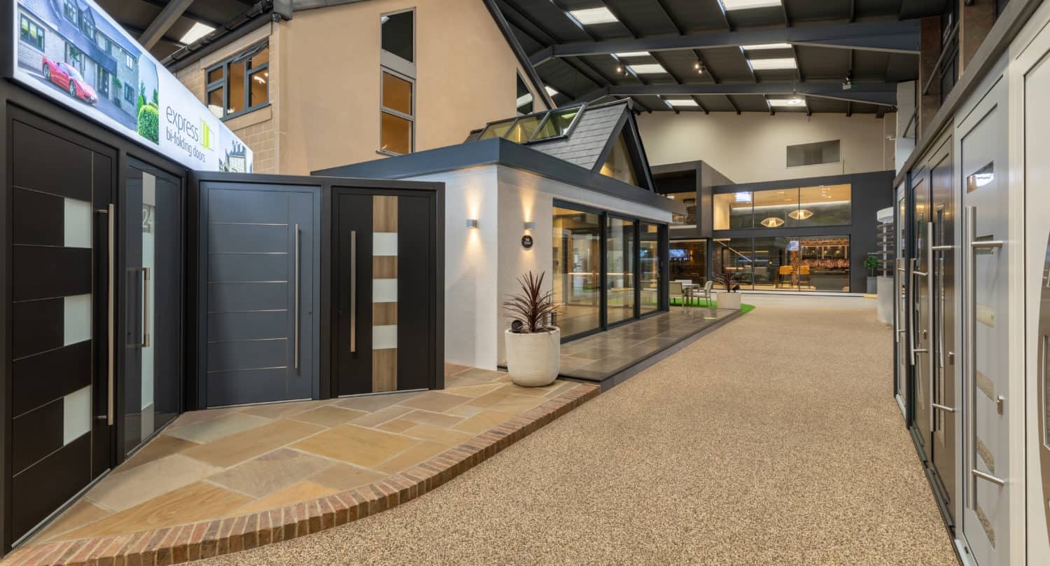 A modern indoor showroom displays various exterior doors and front doors, with a central model house, potted plant, and a spacious layout enhanced by glass roofing under a high ceiling.