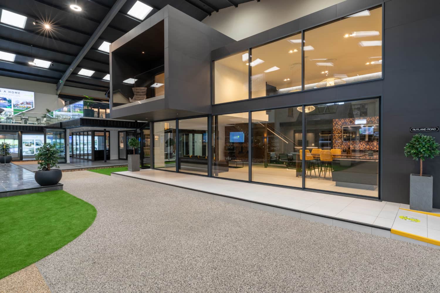 A modern indoor showroom featuring bespoke glass solutions with large windows, a two-story structure, artificial grass, potted plants, and contemporary furnishings—all under a high ceiling with skylights.