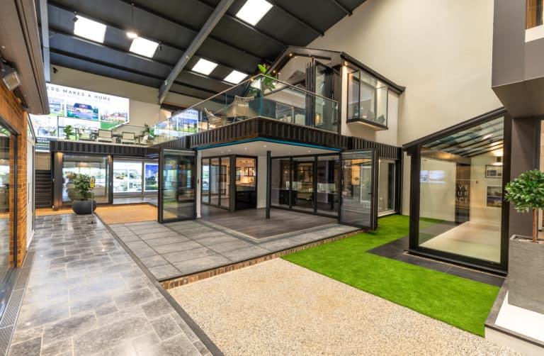A modern showroom with glass roofing, large windows and front doors, a two-story display structure, tiled floors, artificial grass, and bright lighting showcasing home design features.