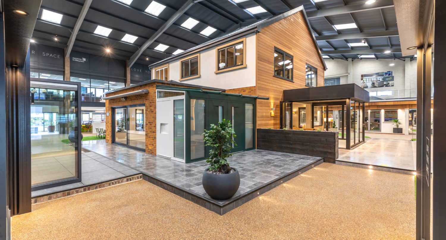 A spacious showroom displays a modern two-story house with large windows, bifold doors, and wood accents. Indoor plants and bright lighting enhance the contemporary, open atmosphere.
