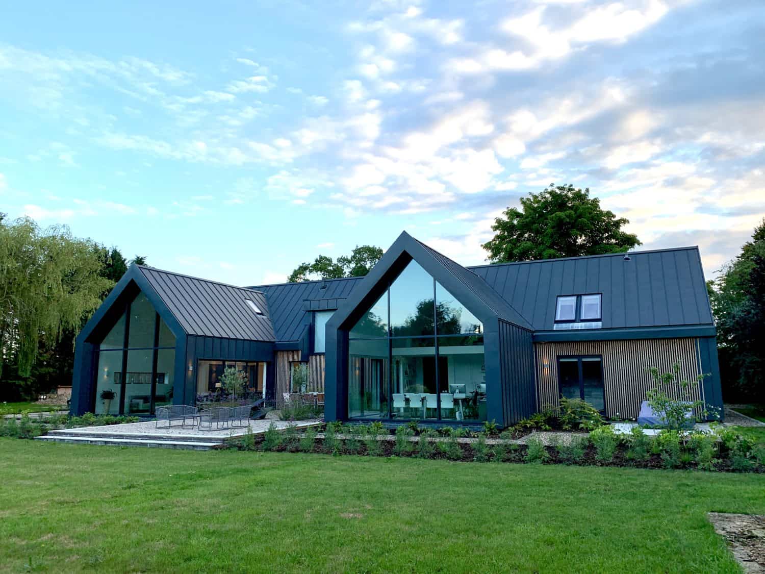 Modern house with large glass windows, dark metal roof, and bespoke glass solutions, surrounded by a grassy lawn and trees under a partly cloudy sky. The home features sharp angles and contemporary architectural design.