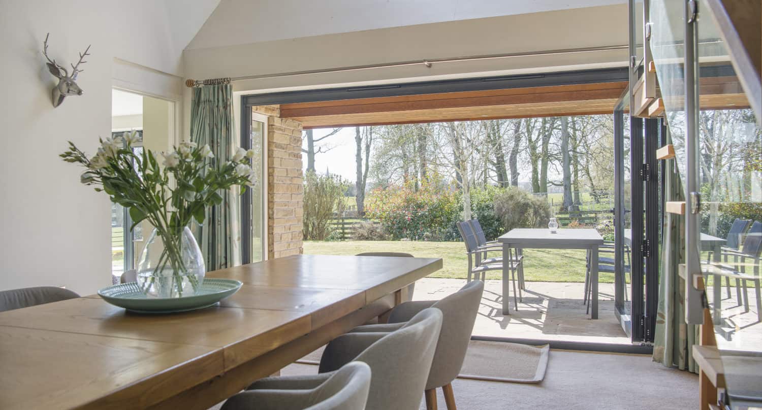 Bright dining room with a wooden table and grey chairs, large bifold doors open to a patio with outdoor seating, and a view of a sunny garden with trees and green grass.