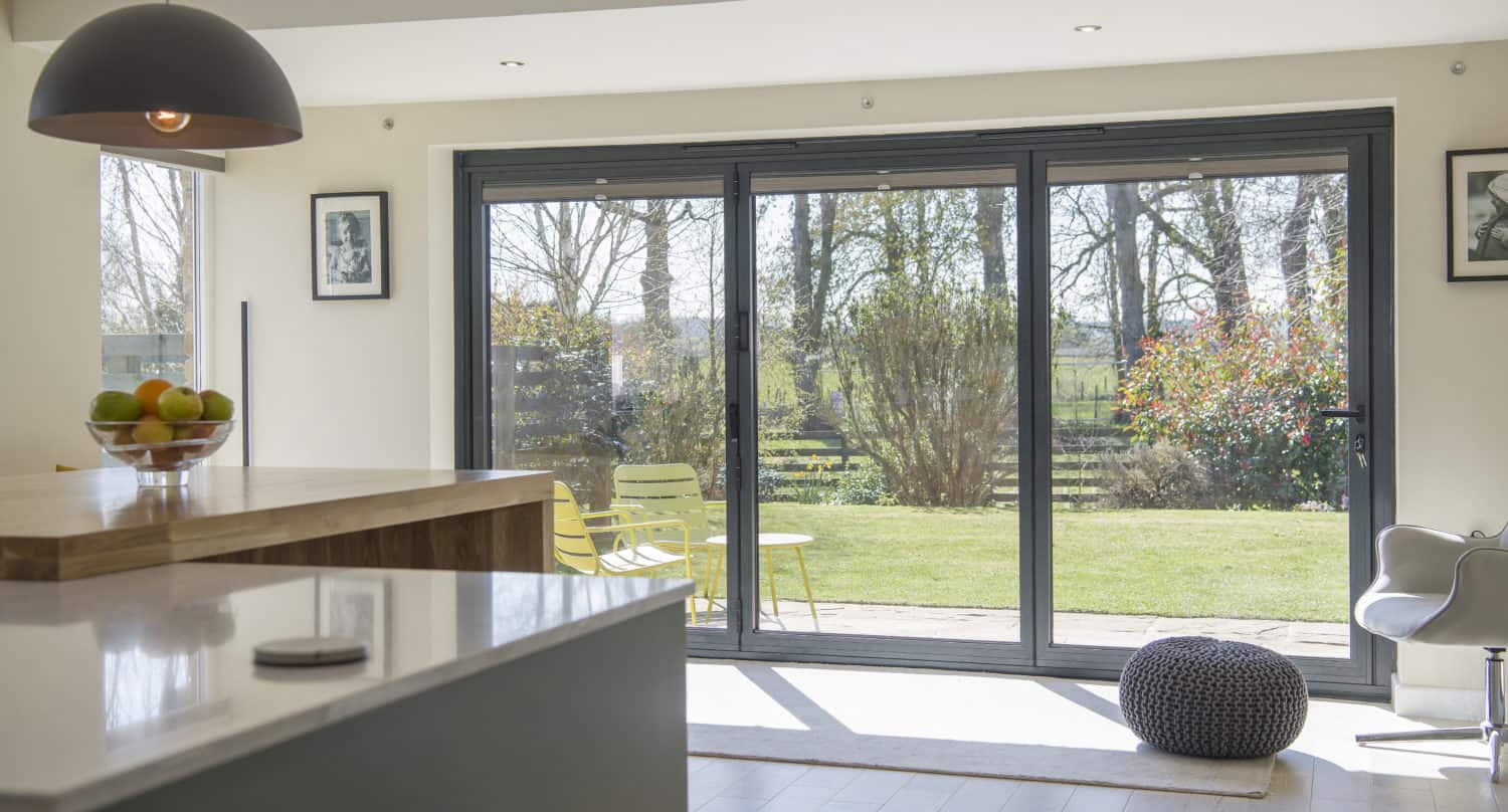 Modern kitchen with bespoke glass solutions—large sliding doors open to a sunny backyard with trees and green grass. A bowl of fruit sits on the counter, and yellow chairs and a table await outside on the patio.