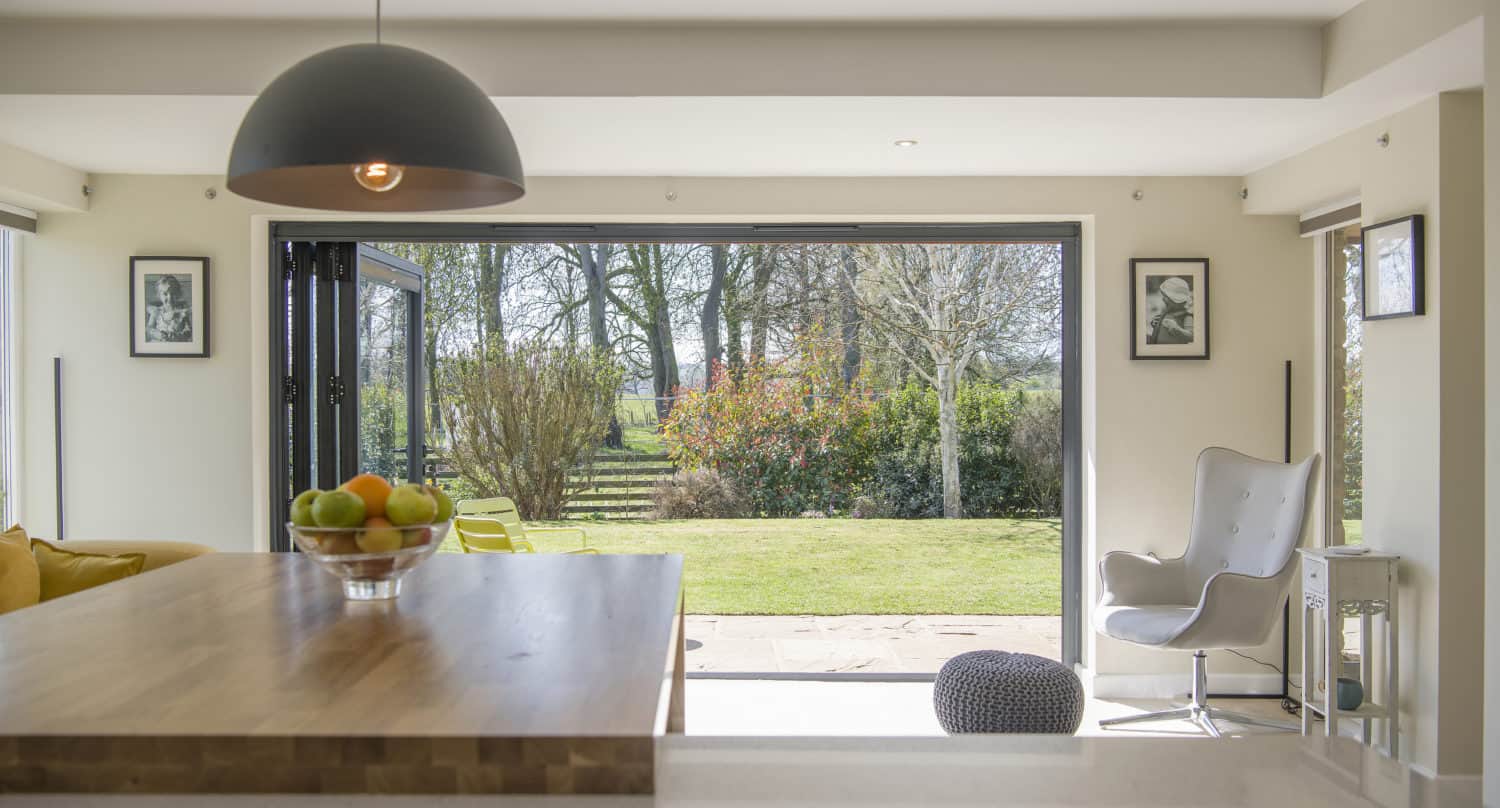Modern kitchen with a wooden island and bowl of fruit in the foreground, opening to a bright living space with large bifold doors revealing a green garden and trees outside. Natural light fills the room.