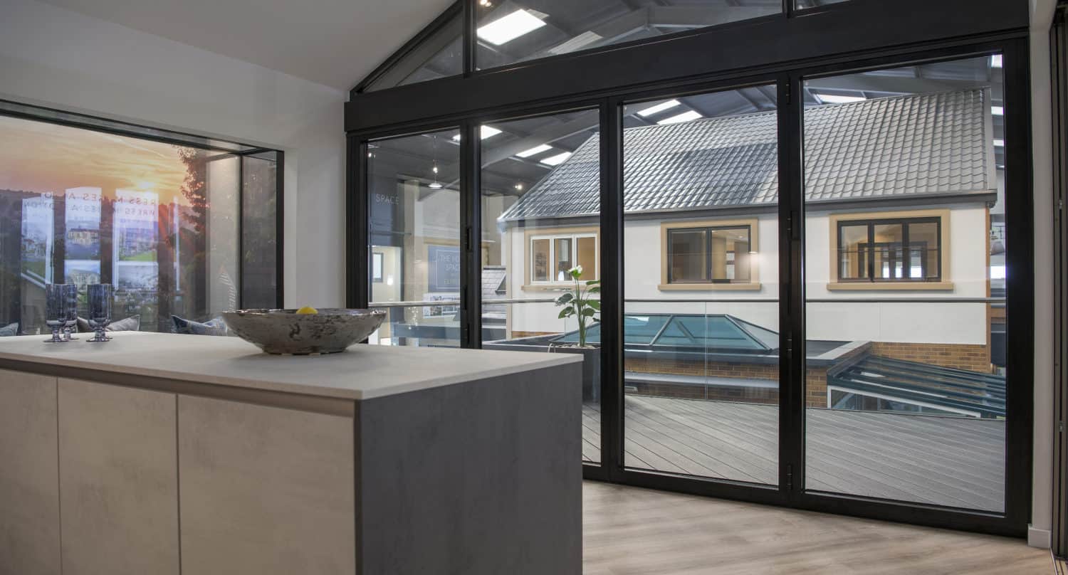 Modern interior space with large glass sliding doors, a light wood floor, and a contemporary kitchen island. The sliding doors open to an enclosed patio with a view of another building and a skylight roof.