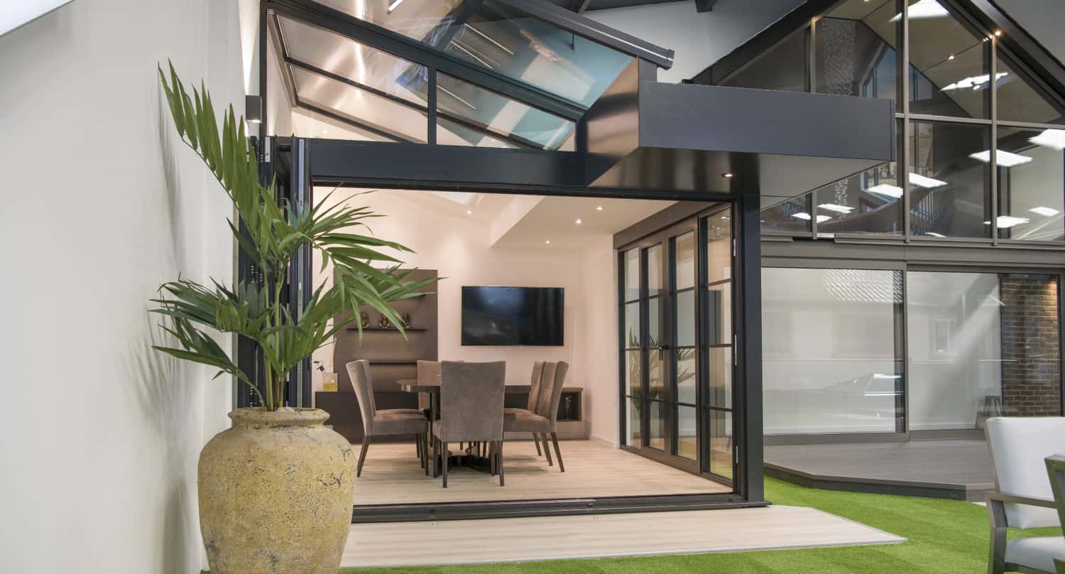 Modern indoor-outdoor space with bespoke glass solutions, including a large sliding door, dining table with chairs inside, a tall potted plant, green artificial grass flooring, and abundant natural light.