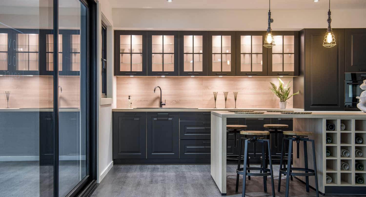 Modern kitchen with dark cabinets, glass-front upper cupboards, a light backsplash, and under-cabinet lighting. An island with stools, pendant lights, and built-in wine storage is in the center. Large sliding doors on the left.