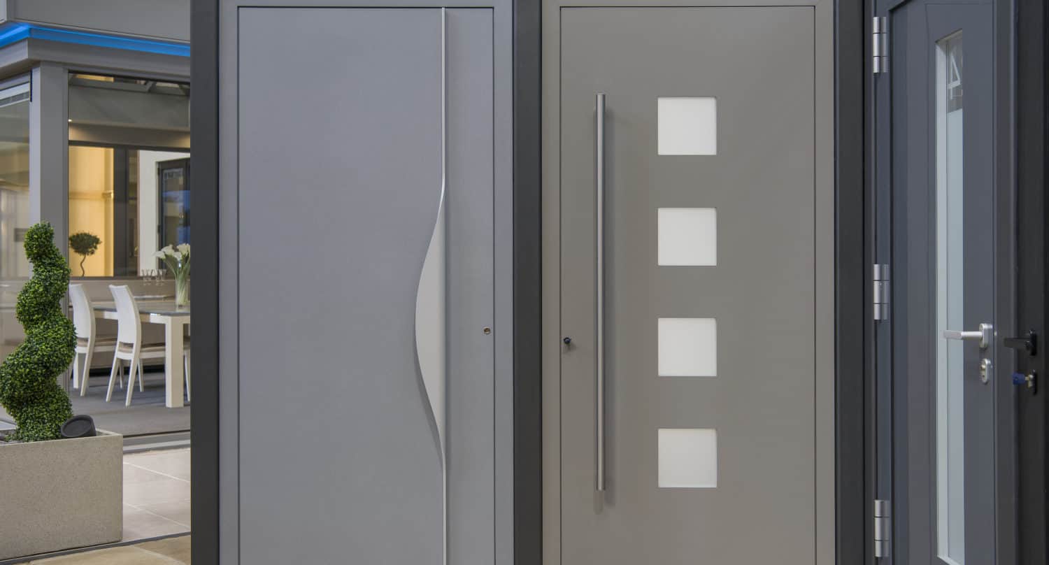 Two modern gray front doors side by side; the left door has a vertical wave pattern, while the right door features four square windows arranged vertically and a long vertical handle, both standing out beneath stylish glass roofing.