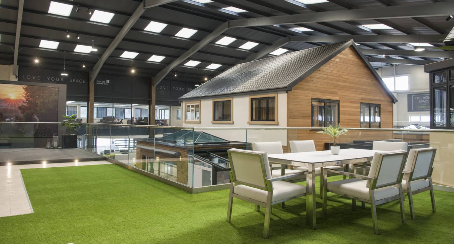 Modern indoor space with artificial grass flooring, a white table and chairs, glass railing, and a house-like structure with wooden siding under a high, metal beamed ceiling with skylights and bespoke glass solutions.