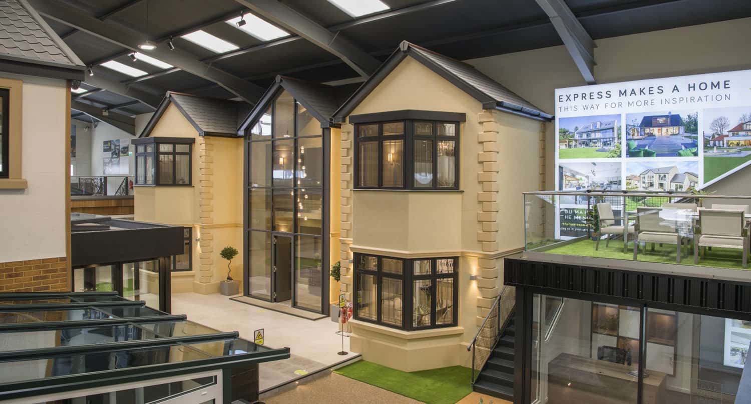 A modern showroom displays full-scale model homes with large windows, beige exteriors, black trim, and bespoke glass solutions like sliding doors. Upstairs, a glass-walled meeting area overlooks a sign reading “Express Makes a Home.”.