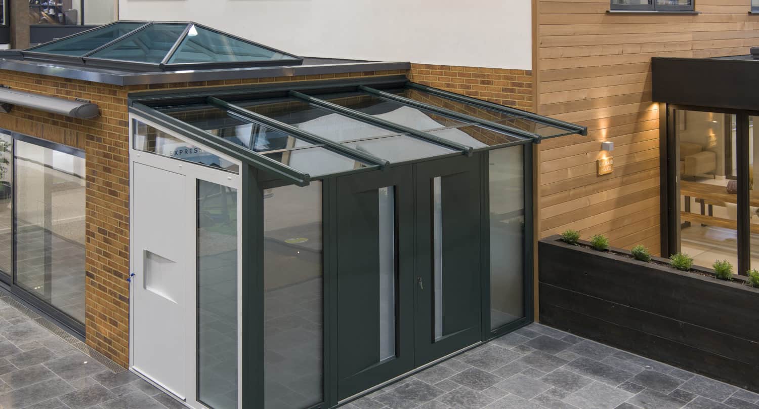 Modern glass and metal entryway extension with large windows and sliding doors, attached to a brick and wood-paneled building, featuring gray tile flooring, glass roofing, and exterior lighting.