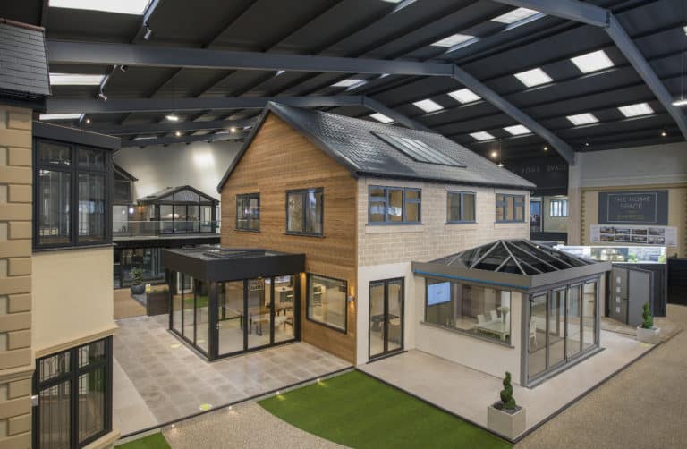 A spacious indoor showroom displays modern house models with large windows, sliding doors, and conservatories under a high, skylit ceiling. The area features artificial grass, bespoke glass solutions, and landscaped walkways.