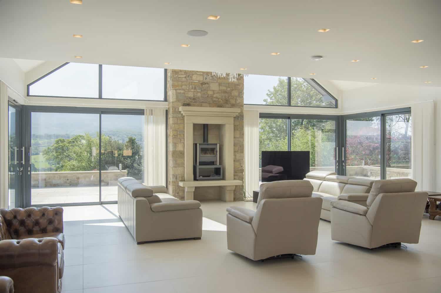 Spacious, modern living room with large windows, beige leather sofas, a stone fireplace, white tile floor, and views of greenery and mountains outside. Sliding doors open to the scenery, while a flat-screen TV sits to the right of the fireplace.