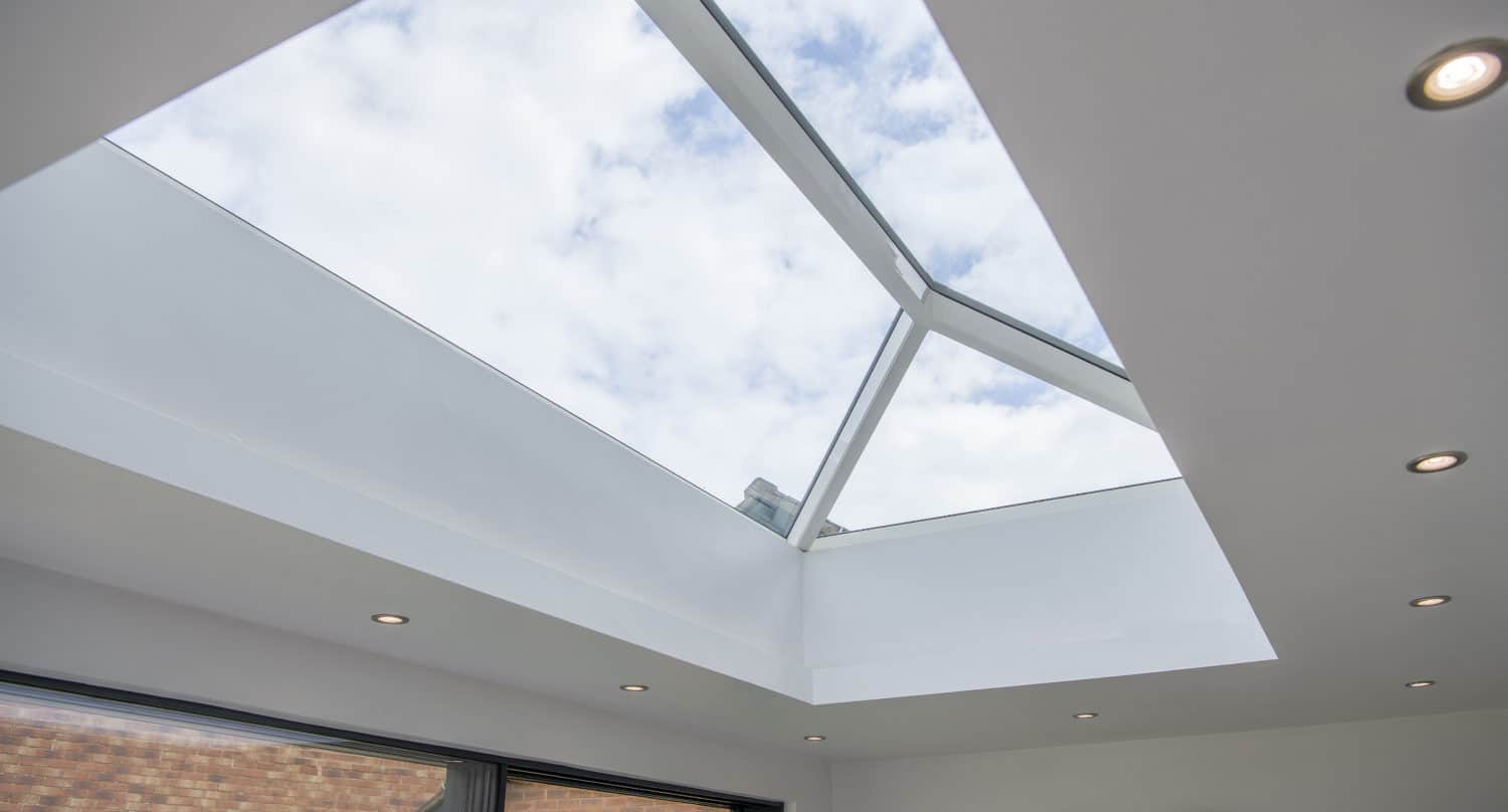 Large skylight window set into a white ceiling, showing a view of a partly cloudy sky; recessed ceiling lights and sleek glass roofing elements complement the modern architectural design.