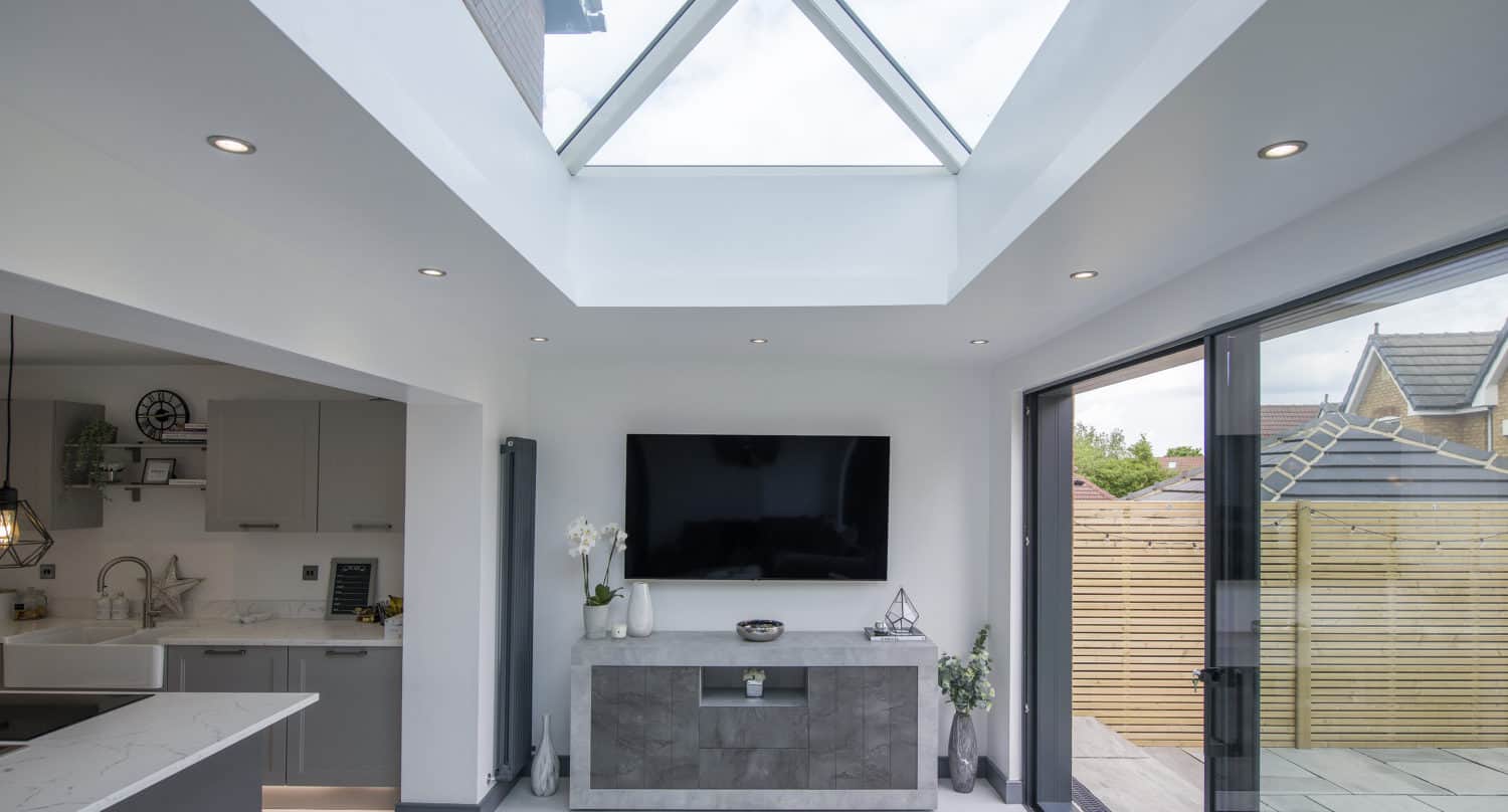 Modern, bright living space with a large skylight and sleek cabinetry. Glass bifold doors open to a patio with wooden fencing, while the adjacent kitchen area is visible on the left. Wall-mounted TV completes the look.