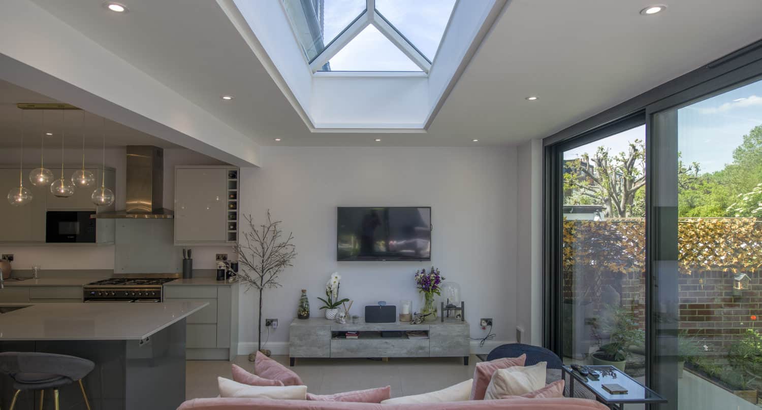 Modern living room with a skylight, pink sofa, wall-mounted TV, sliding doors opening to a garden, and a kitchen area with bar stools and pendant lights. The bright, airy space features minimal decor for a fresh feel.