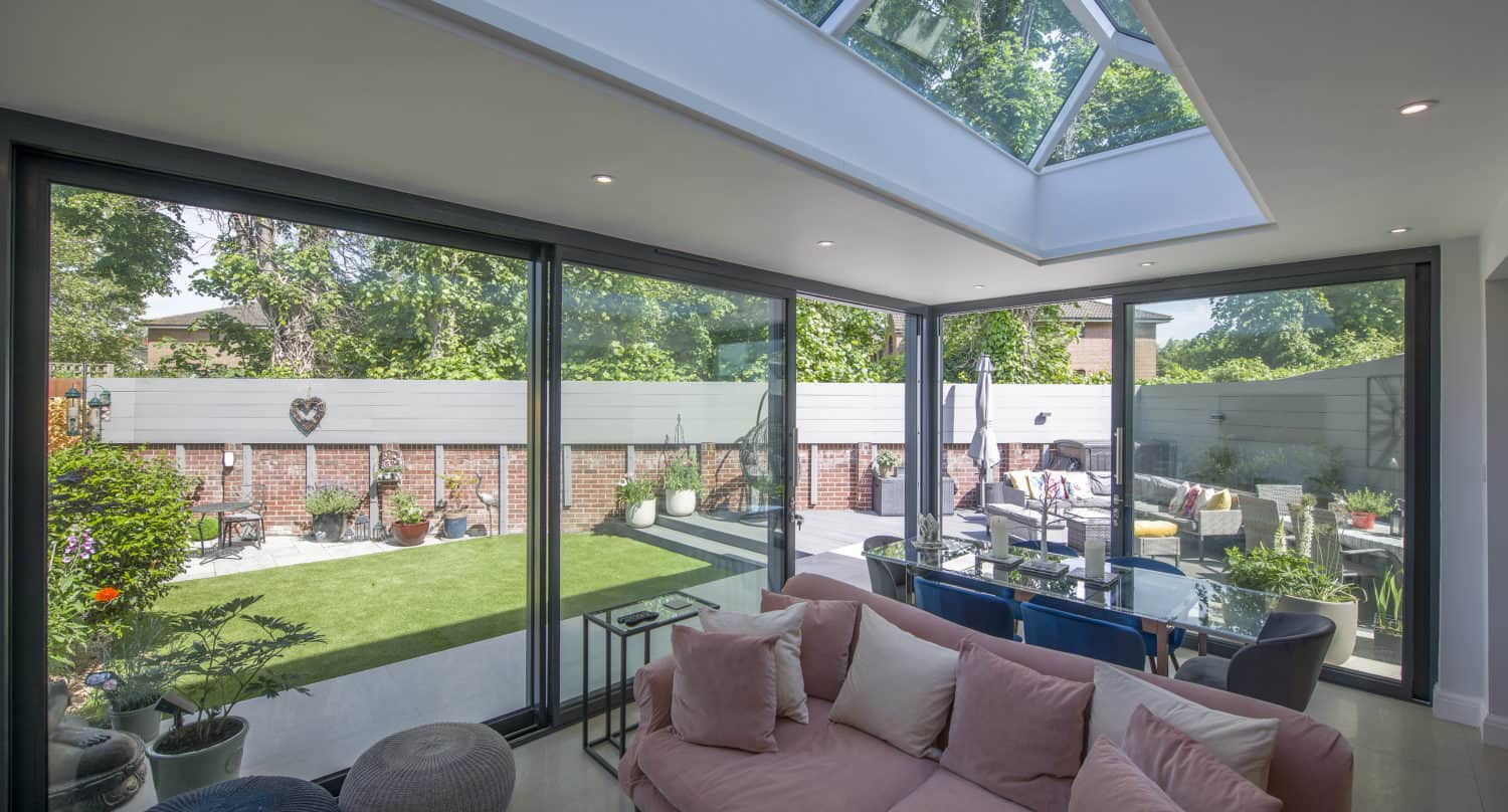 A modern living room with a pink sofa and large sliding doors opens to a sunny, landscaped backyard with patio seating, potted plants, and bespoke glass solutions like a skylight overhead that fills the space with natural light.
