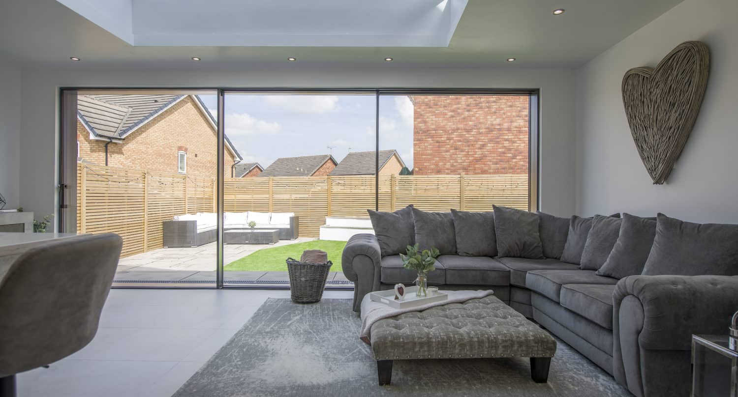Modern living room with large gray sectional sofa, ottoman, and rug. Bespoke glass sliding doors open to a fenced backyard. Skylight above lets in natural light. Wicker heart decor hangs on the white wall.