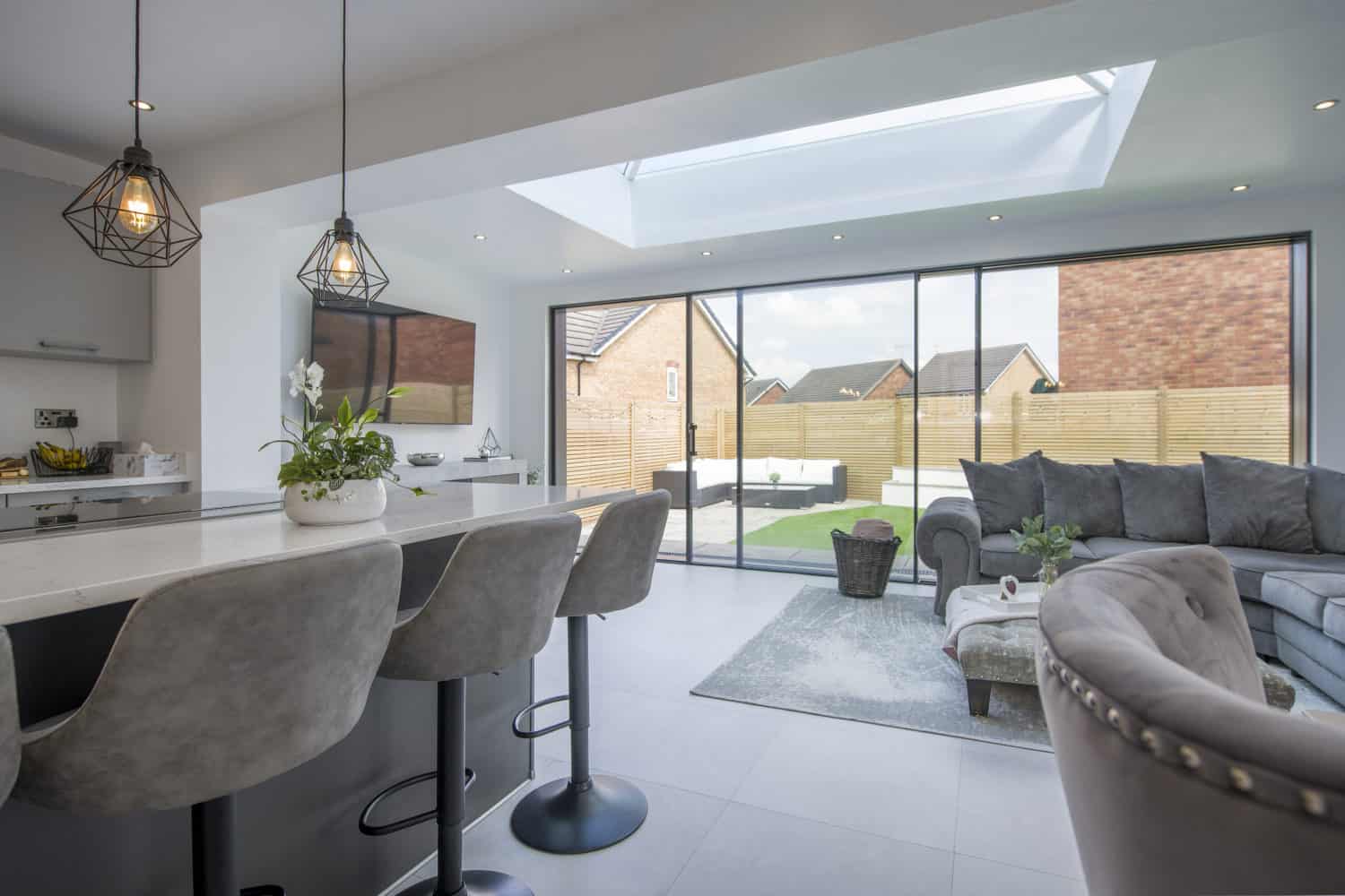 Modern open-plan kitchen and living room with grey bar stools, pendant lights, a glass roofing skylight, and sliding glass doors opening to a backyard with lawn and fencing. A grey sofa and coffee table are in the living area.