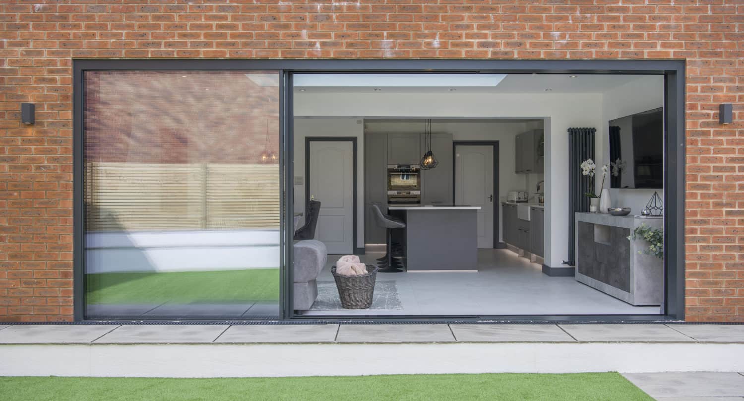 Open sliding bifold doors reveal a modern kitchen and living area with neutral tones, an island, and contemporary décor, viewed from an outdoor space with artificial grass and a brick exterior wall.