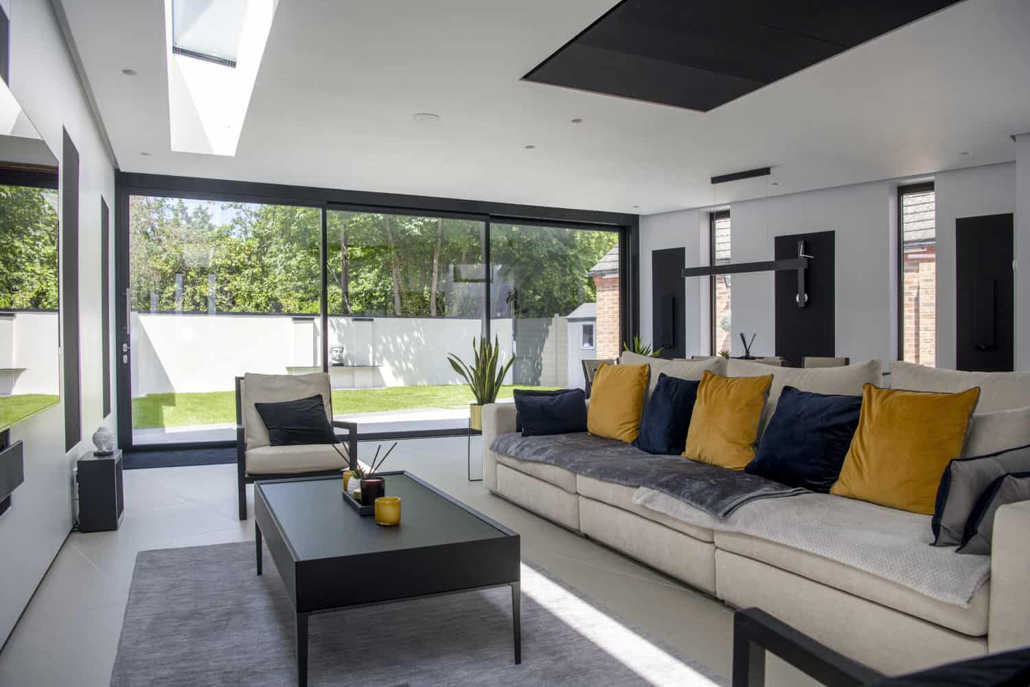 A modern living room with a beige sectional sofa decorated with blue and yellow cushions, a black coffee table, large floor-to-ceiling windows, and bespoke glass solutions offering a view of the grassy backyard and trees outside.