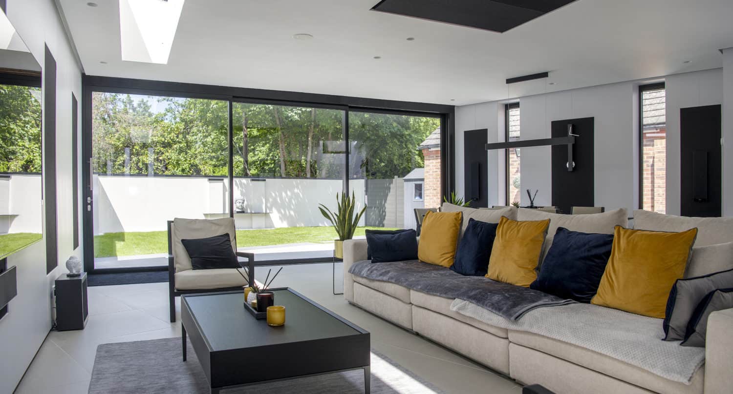 A modern living room with a beige sectional sofa decorated with blue and yellow cushions, a black coffee table, large floor-to-ceiling windows, and bespoke glass solutions offering a view of the grassy backyard and trees outside.