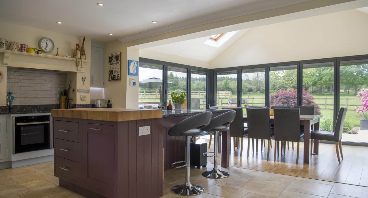 Modern kitchen with a wood island, bar stools, and an open dining area featuring bifold doors and large windows offering views of a green garden and trees outside. Ample natural light fills the space.