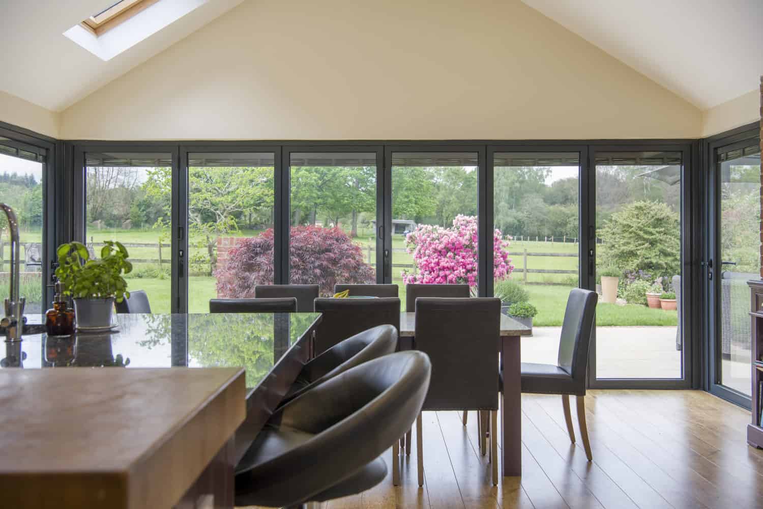 Modern dining area with black chairs and a glass table, featuring large bifold doors opening to a patio and garden with colorful bushes and a green lawn, filling the space with abundant natural light.