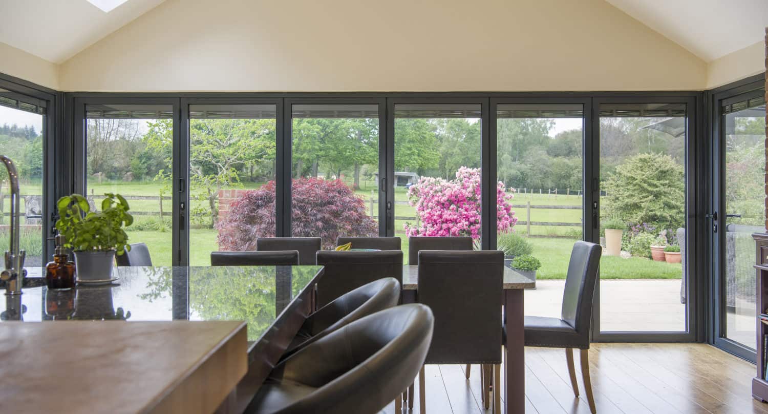 Modern dining area with black chairs and a glass table, featuring large bifold doors opening to a patio and garden with colorful bushes and a green lawn, filling the space with abundant natural light.