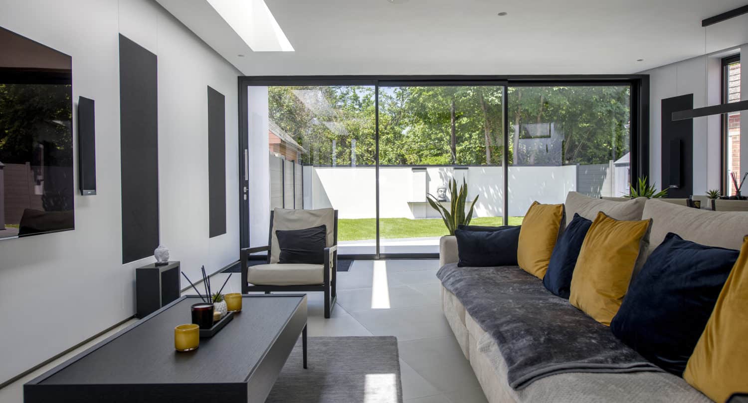 A modern living room with a gray sofa and black and yellow cushions, a black coffee table, a chair, and sliding doors opening to a sunny garden with a white fence and green trees.