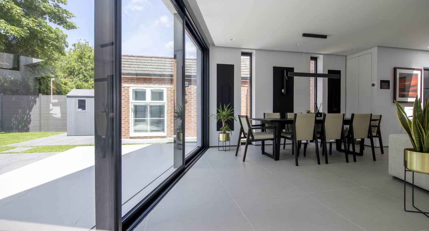 Modern dining area with floor-to-ceiling sliding doors, letting in natural light. The room features a dining table with chairs, potted plants, light tile flooring, and views of a green backyard with a brick outbuilding.