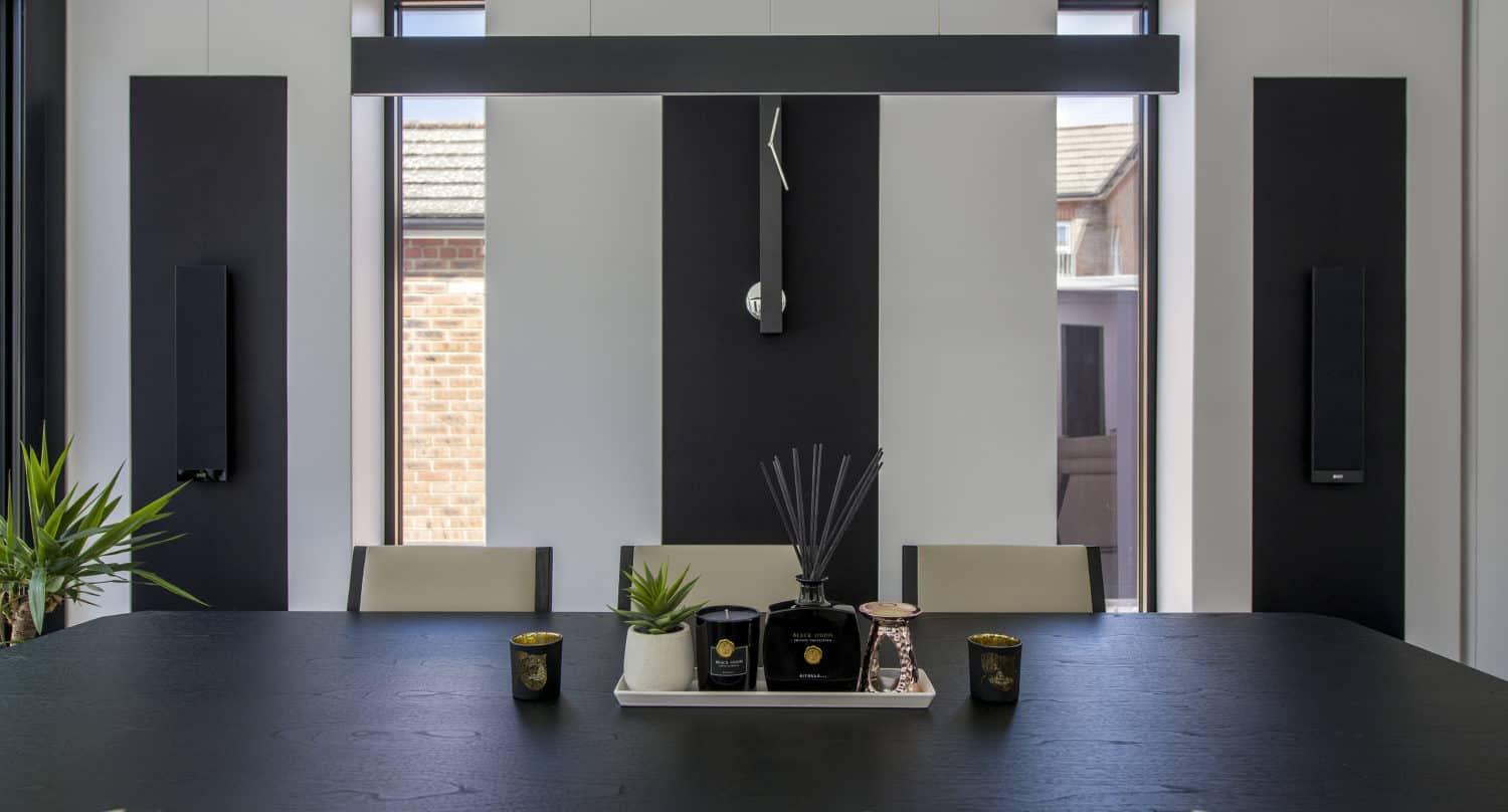 Modern dining room with a black table, cream chairs, decorative plants, candles, and reed diffusers. Large windows and sliding doors let in natural light, while the room features a minimalist black and white color scheme.