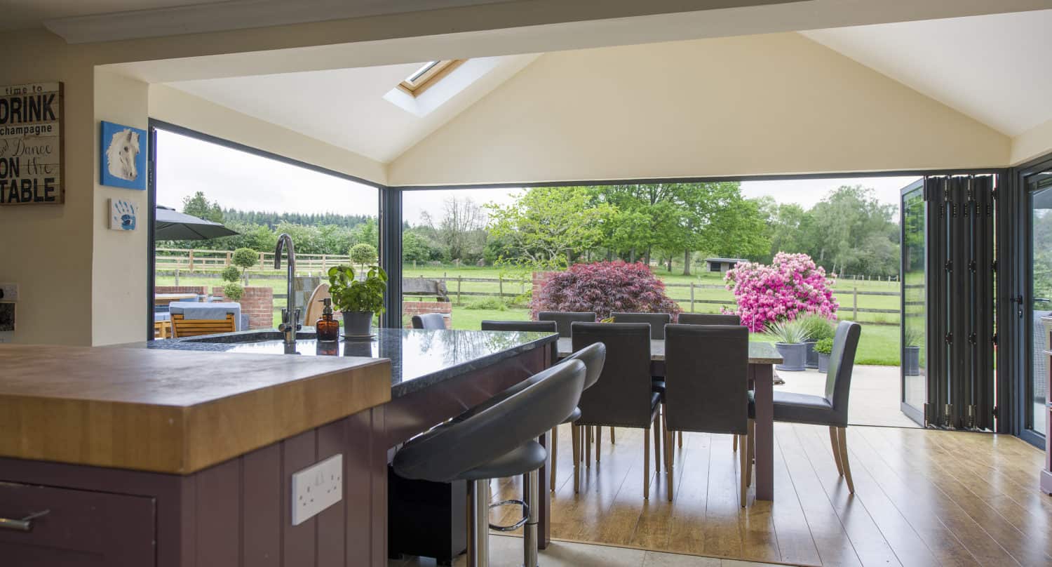 Modern open-plan kitchen and dining area with bifold doors leading to a patio and garden, featuring a large island, barstools, and views of vibrant greenery and pink flowering bushes outside.