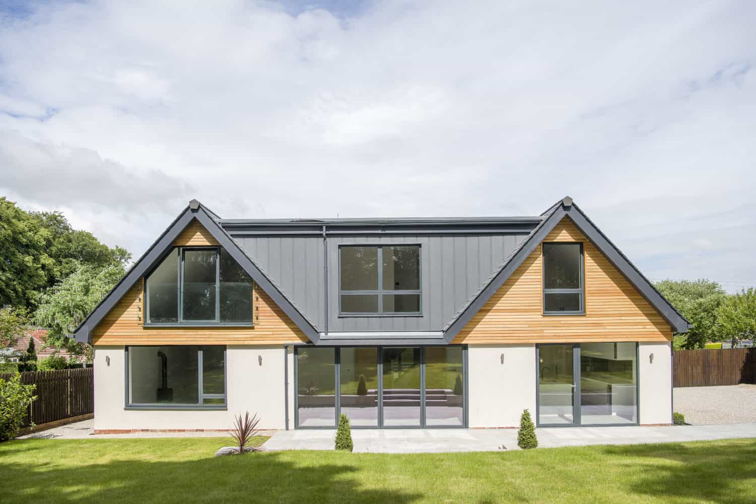 Modern two-story house with large windows, wood and white exterior, a metal roof, and sliding doors opening to a neatly manicured lawn in front. Trees and a wooden fence border the yard.