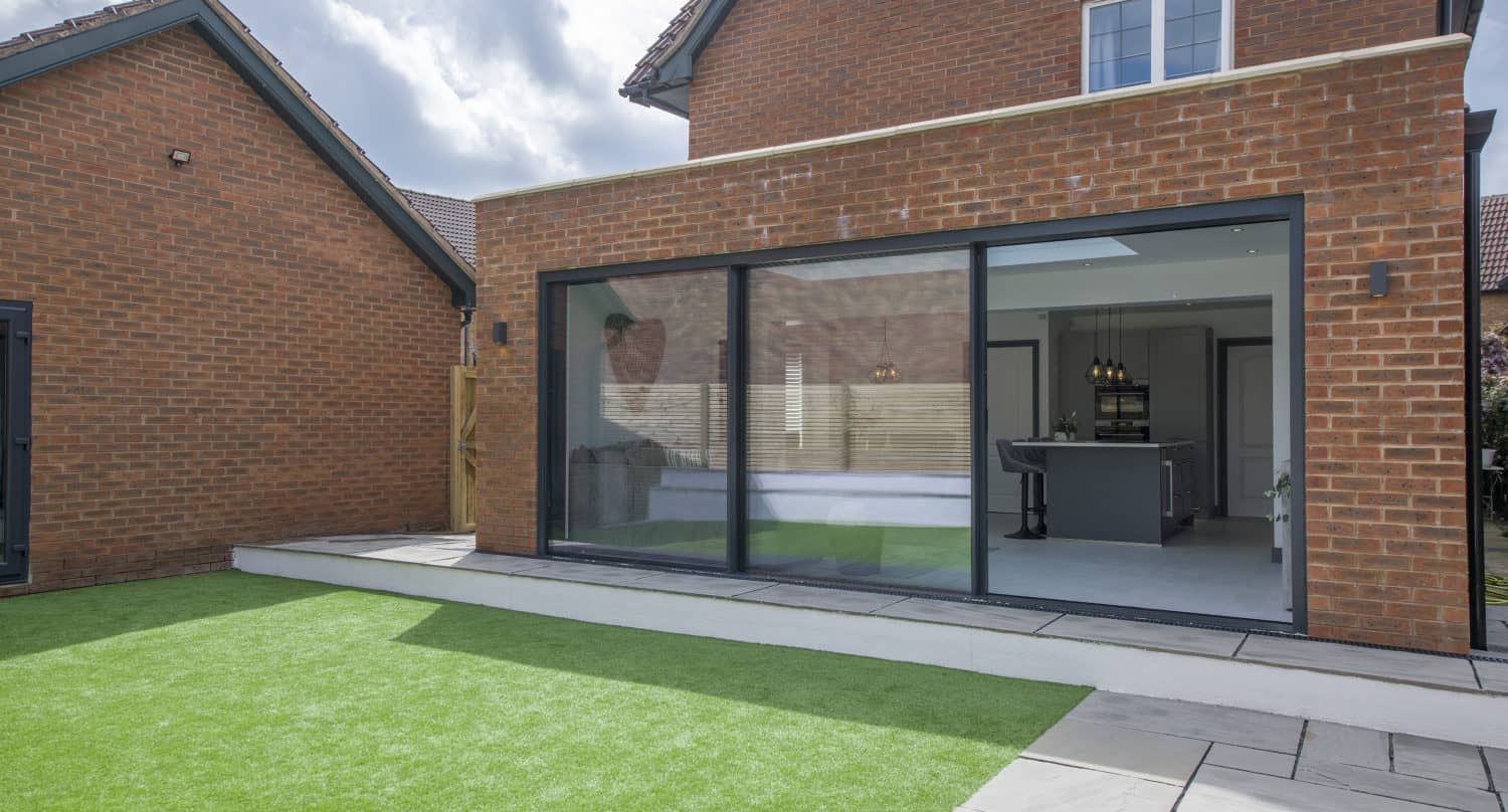 Modern brick house with large sliding doors opening to a backyard featuring artificial grass and a tiled patio area. The kitchen and dining area are visible through the doors. The sky is partly cloudy.