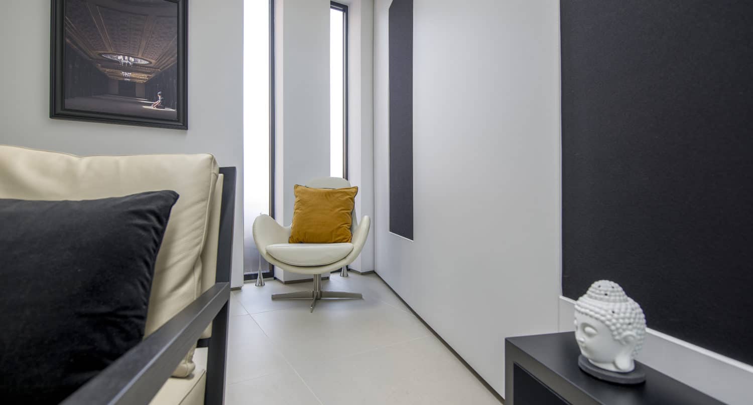 Modern minimalist room with a white armchair, mustard yellow pillow, black pillow on a beige sofa, Buddha head sculpture on a black table, wall art, and tall bifold doors letting in natural light.