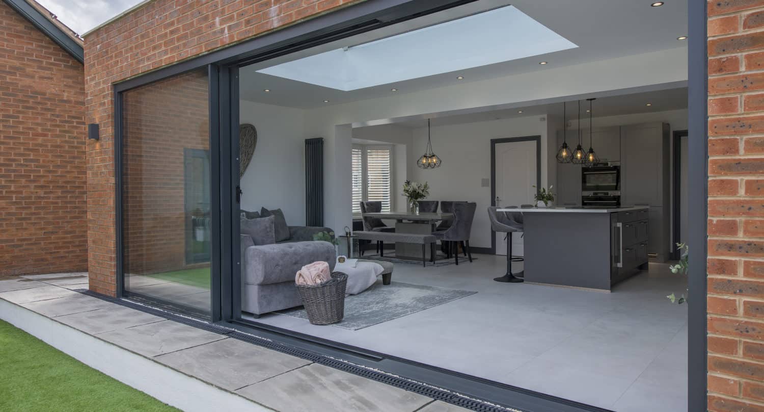Modern open-plan living space viewed through large bifold doors, featuring a grey sofa, dining area, and sleek kitchen, with brick exterior and artificial grass outside.
