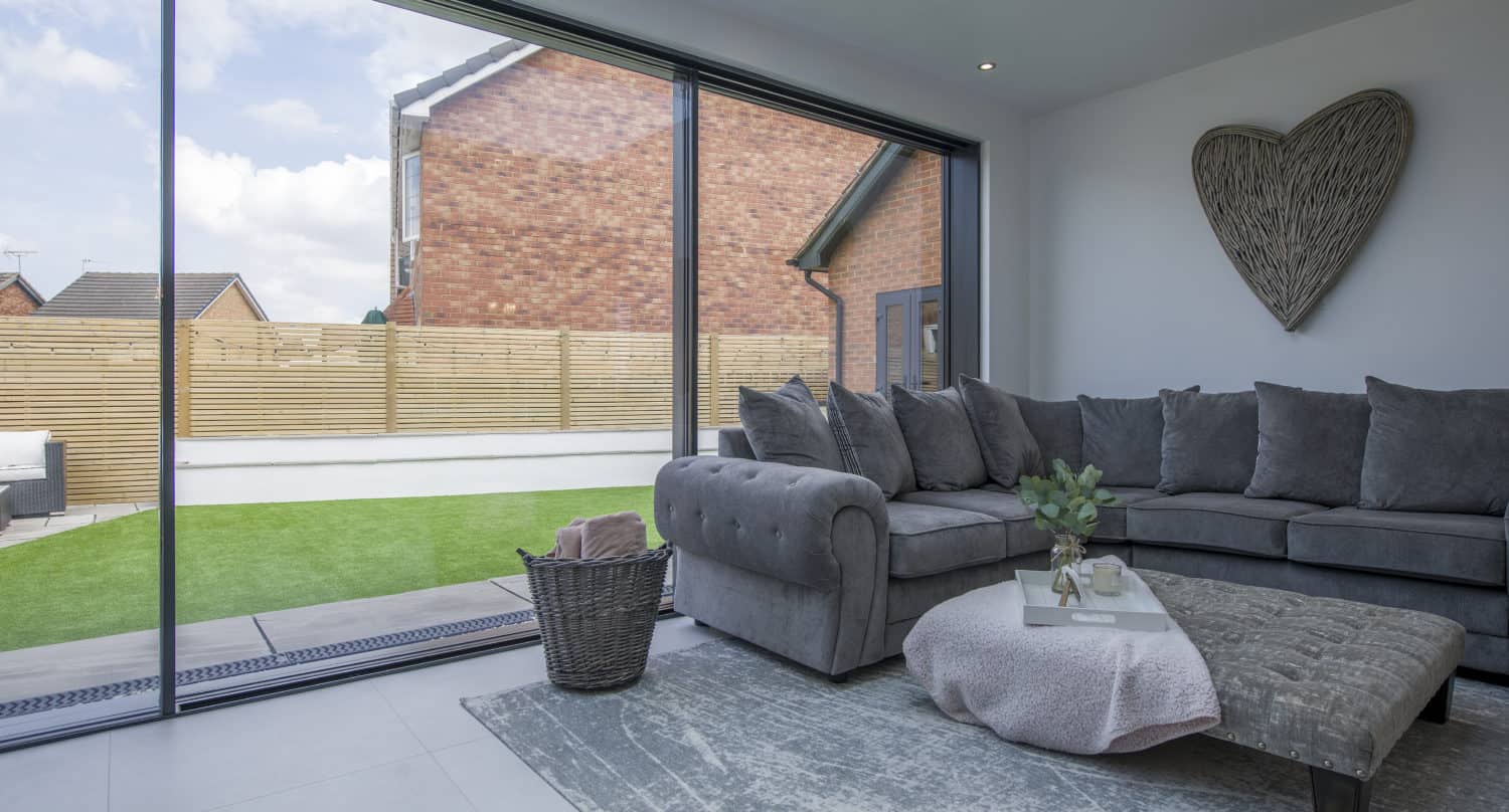 Modern living room with large bifold doors overlooking a backyard with artificial grass. Inside, there’s a gray sectional sofa, a textured rug, a coffee table, and a heart-shaped wall decoration.