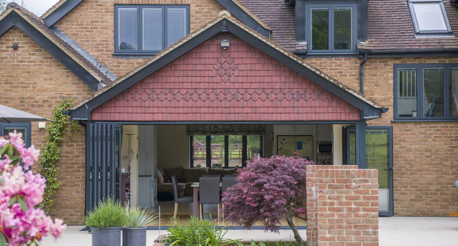 A modern brick house with large bifold doors opening to a spacious interior. The garden features green grass, potted plants, a small red-leafed tree, and blooming pink flowers in the foreground.