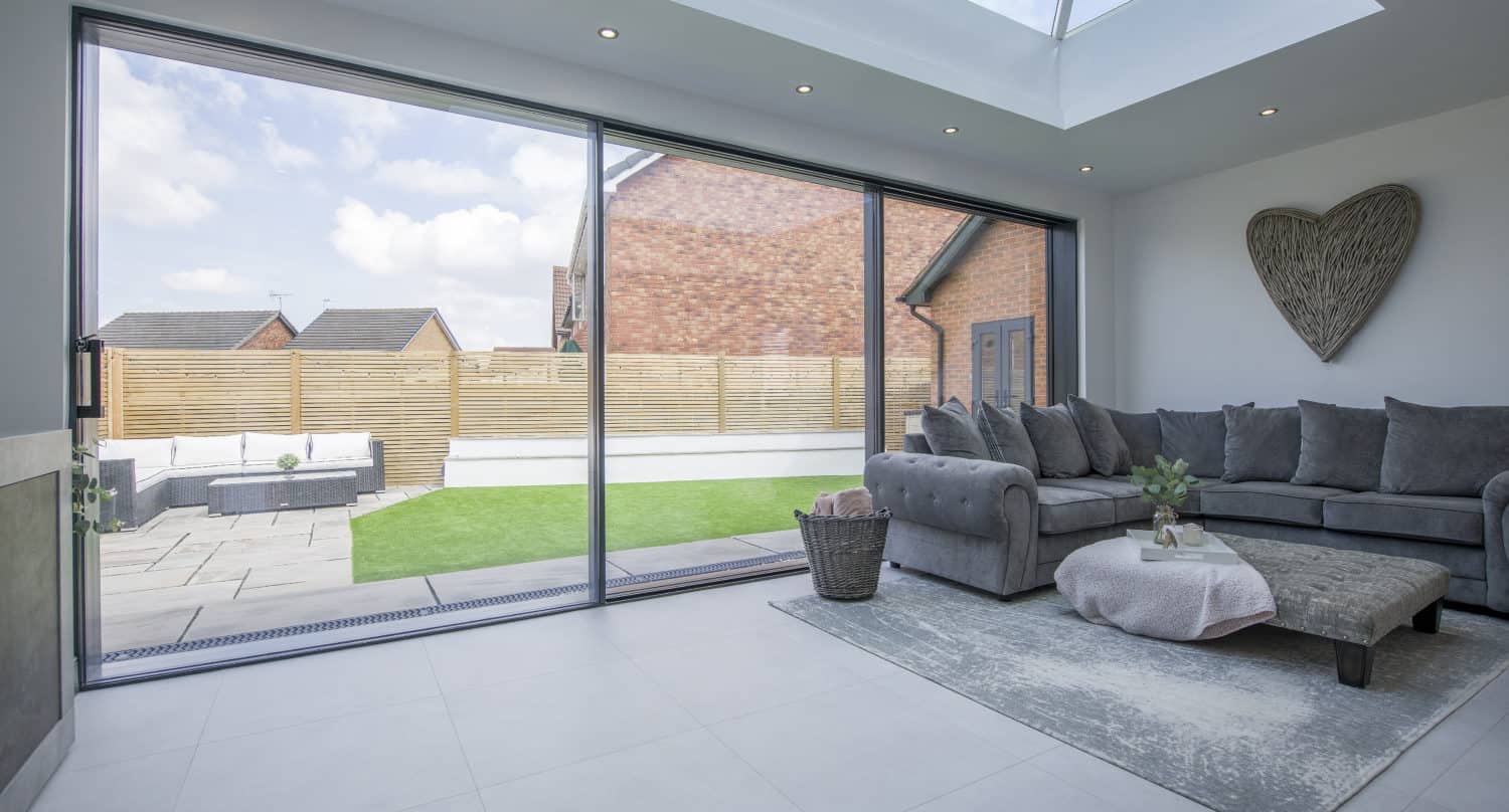 Modern living room with large bifold doors opening to a backyard with lawn and patio. Gray sectional sofa, round coffee table, light rug, and heart-shaped wall decor create a cozy, bright atmosphere.