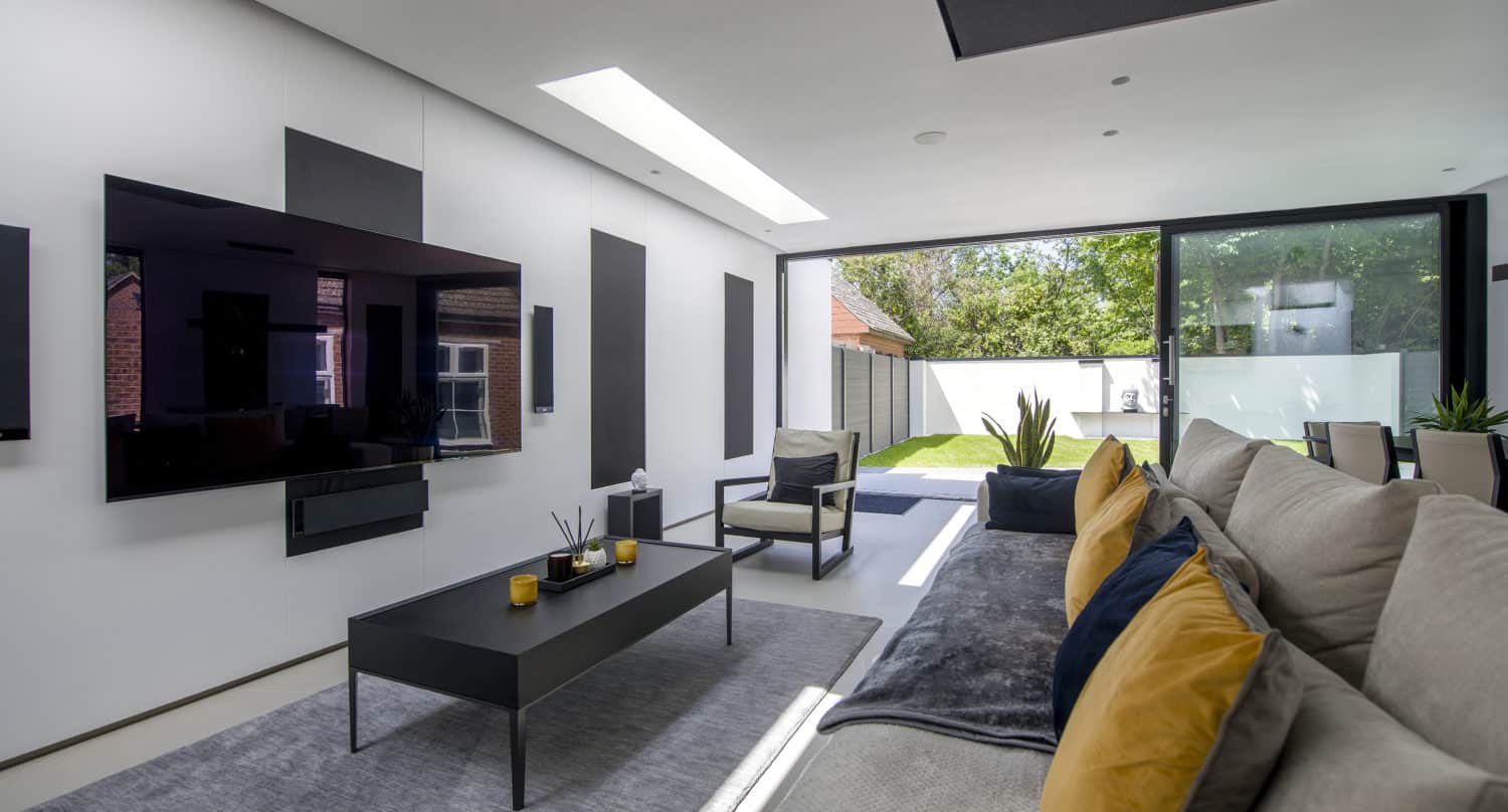 Modern living room with a large wall-mounted TV, sleek black coffee table, gray sofa with yellow pillows, contemporary décor, and floor-to-ceiling windows featuring bespoke glass solutions that overlook a green backyard.