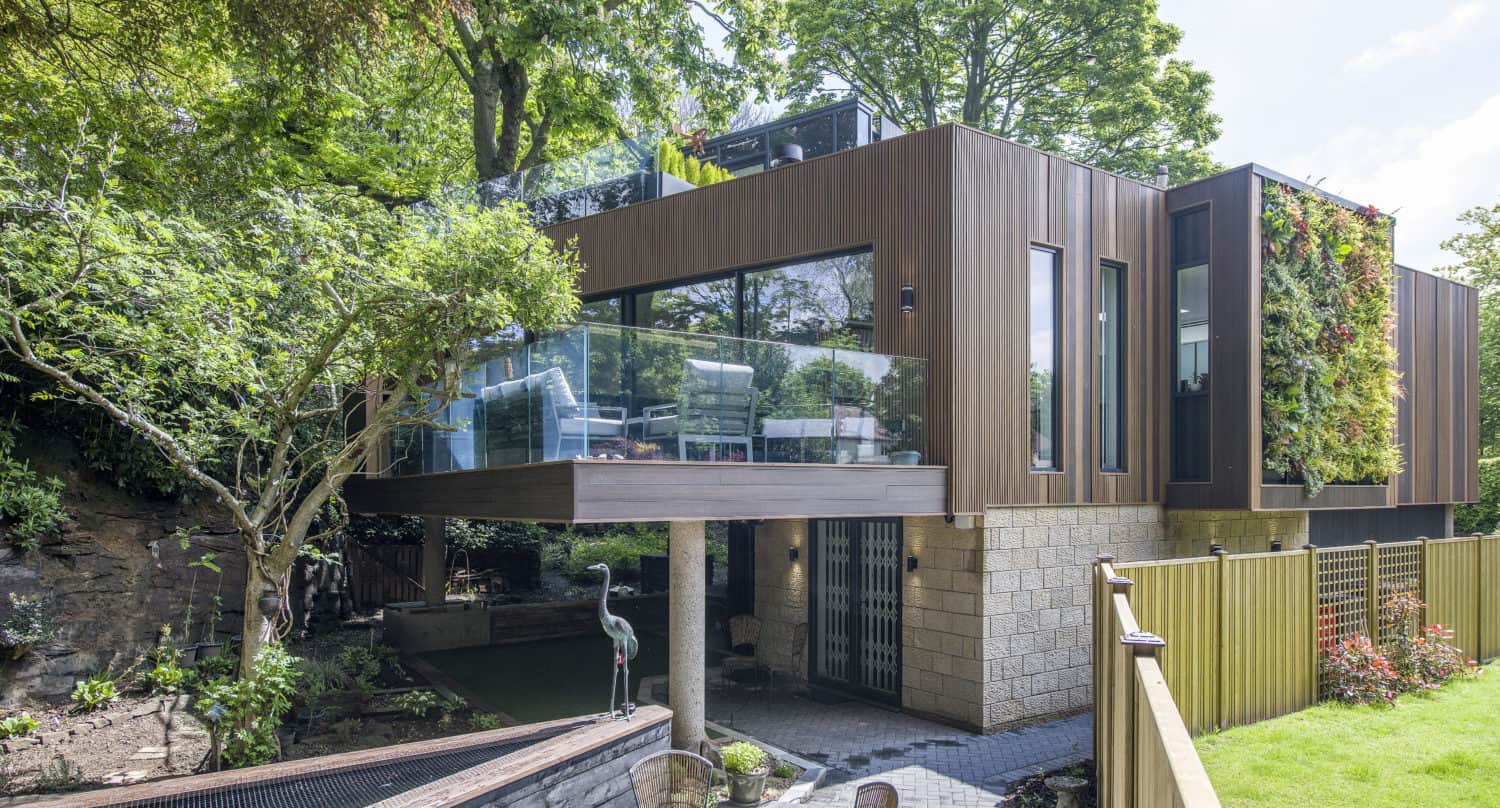 Modern house with large glass windows, wood paneling, and sliding doors, elevated on stone columns. Lush greenery surrounds the home, a vertical garden accents one wall, and a fenced patio area with outdoor furniture sits below.