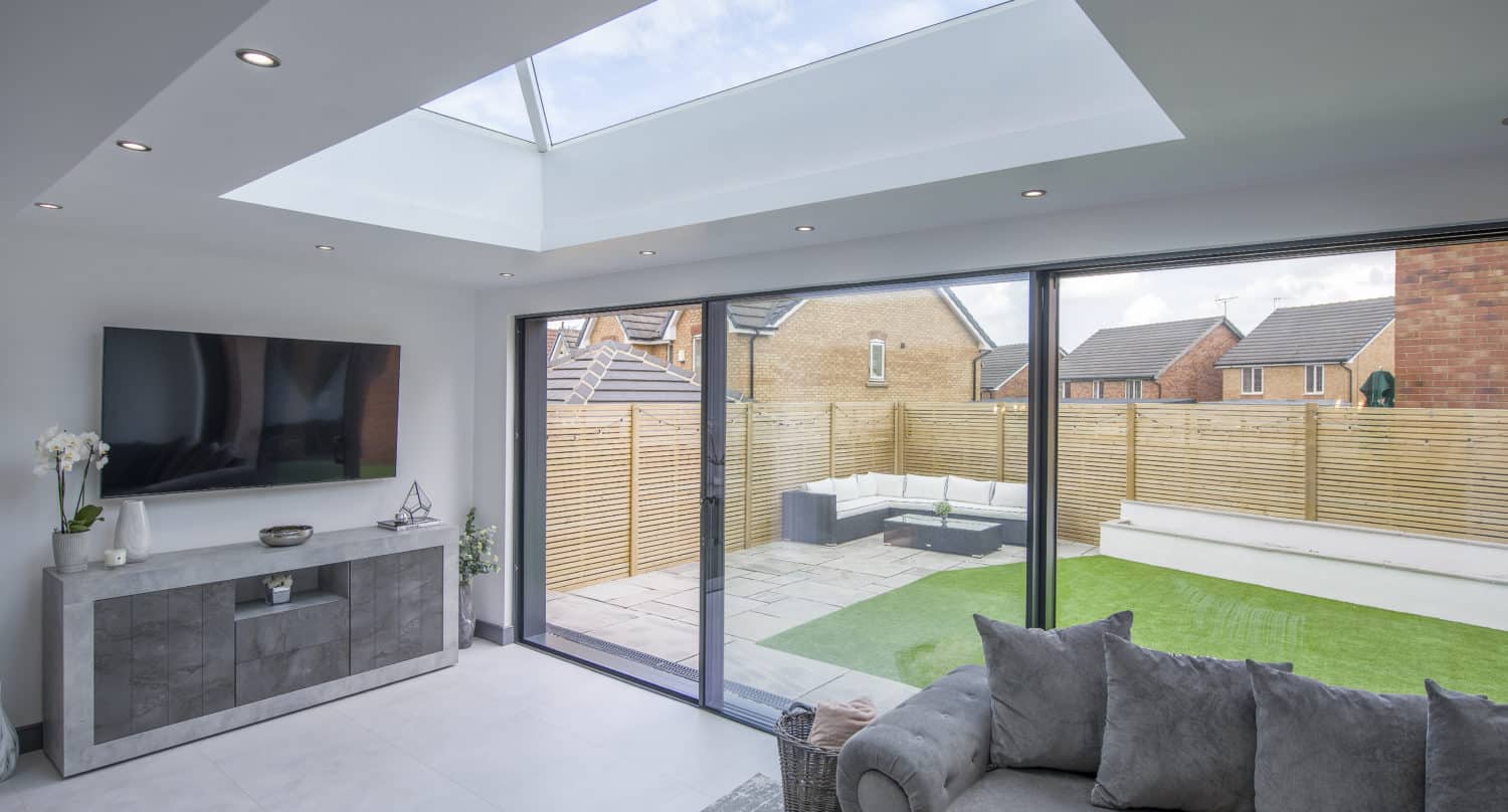 Modern living room with large glass roofing skylight, gray sofa, TV on a console, and sliding glass doors opening to a patio with seating, artificial grass, and wooden fencing. Bespoke glass solutions highlight views of houses beyond the fence.