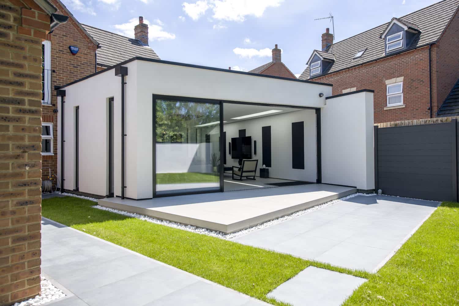 Modern white extension with large glass sliding doors and bespoke glass solutions, flat roof, and minimalist design connected to a brick house. The patio features grey tiles, neatly trimmed grass, and a black fence.