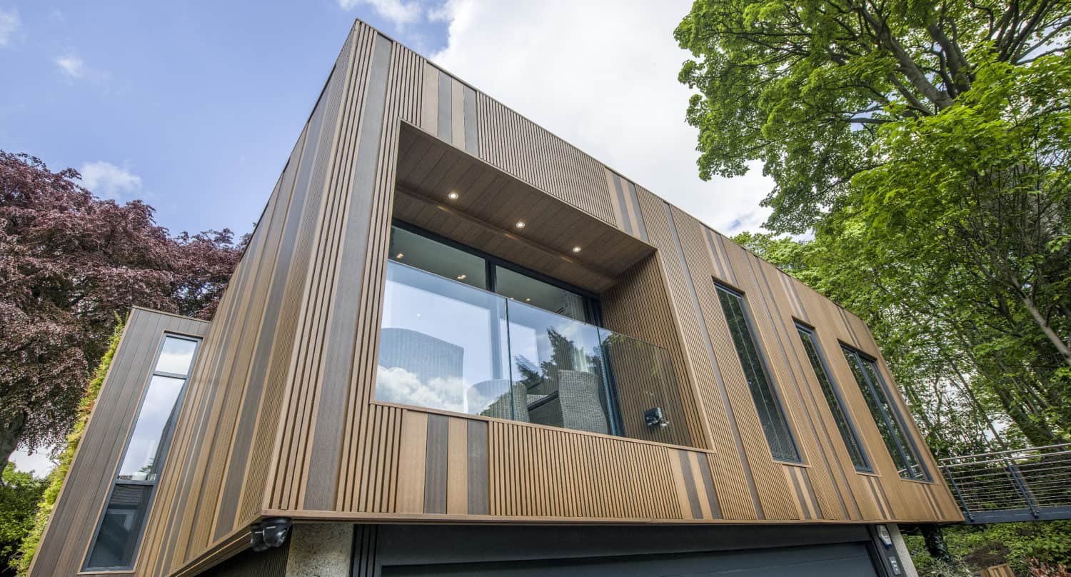 A modern house with a wooden exterior, large windows, and bifold doors opening to a glass balcony, surrounded by tall green trees and set above a gray garage. The sky is partly cloudy.